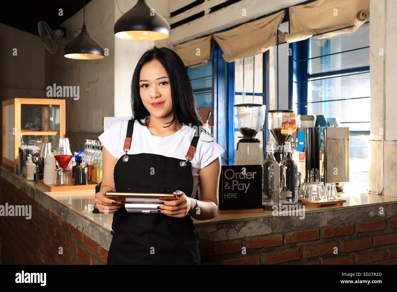 Asian Woman Barista Wearing Black Apron Looking at Camera and Holding Tablet, Standing Next to Cafe Bar Stock Photo