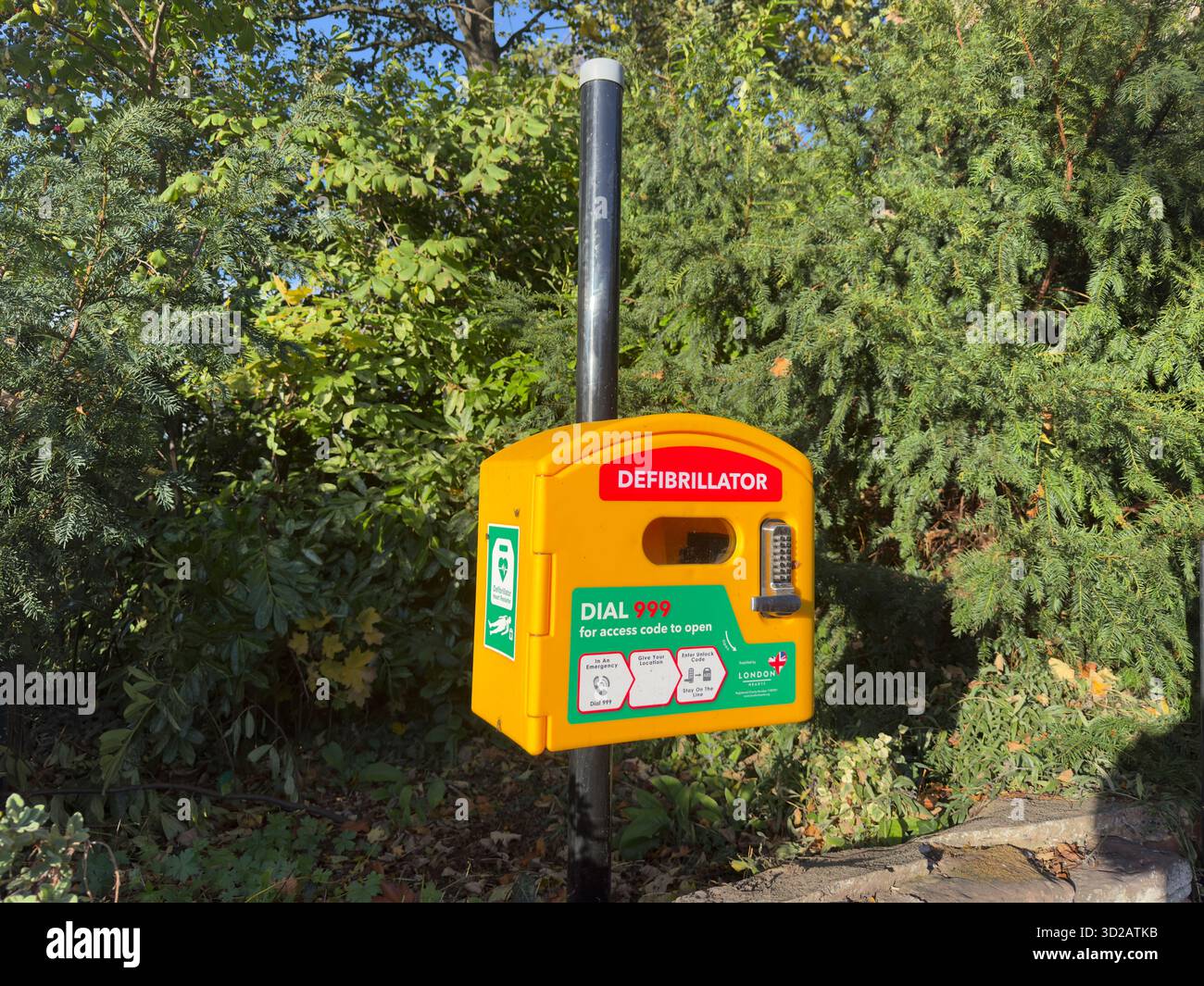 Landscape image of a defibrillator in a secure yellow cabinet fixed to a black metal pole located in front of trees and bushes outside a church. - Smartphone Captured Stock Image