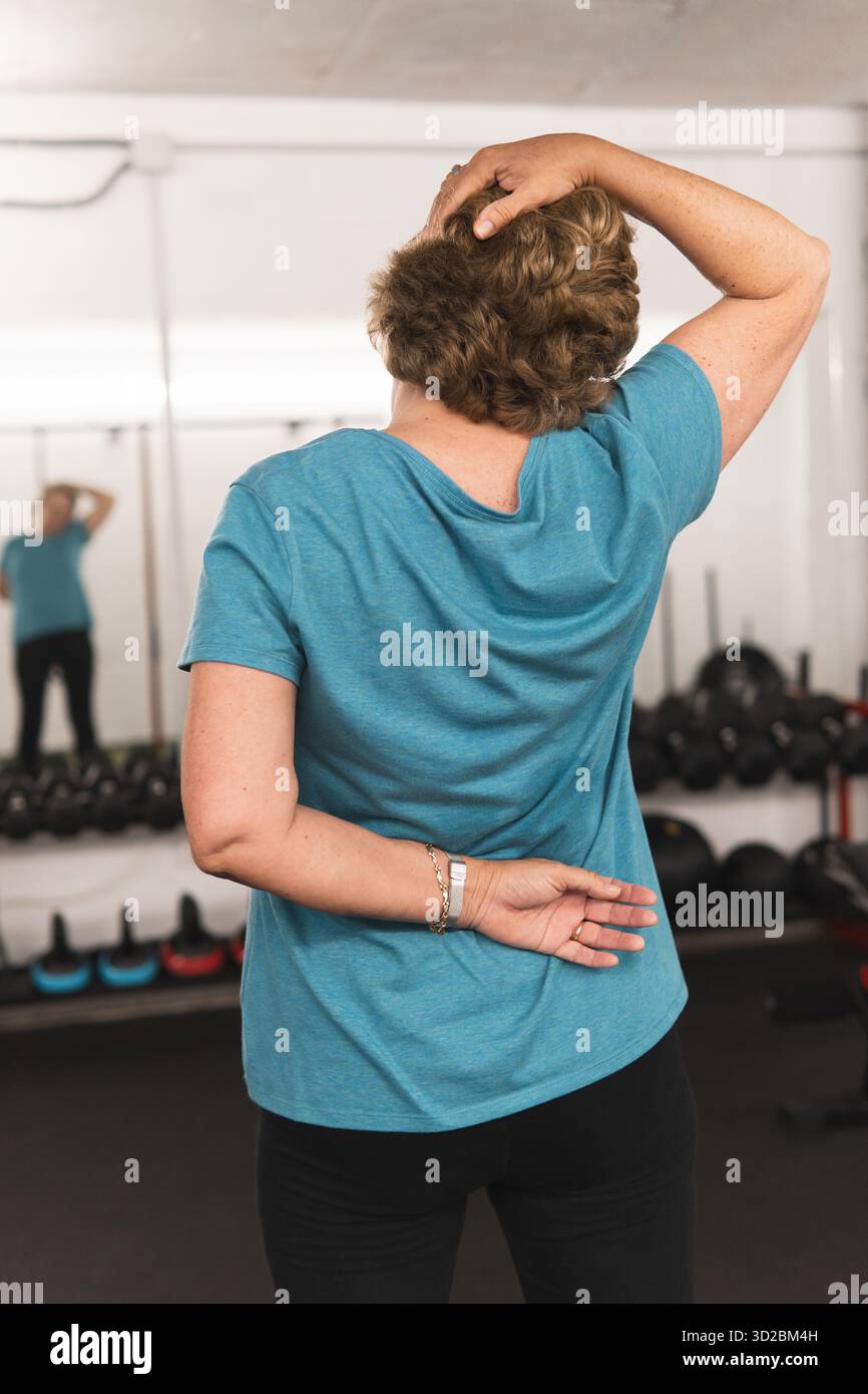 A woman is in a gym, wearing a blue shirt and black pants. She is bending over and touching her head with her hand. Concept of determination and focus Stock Photo