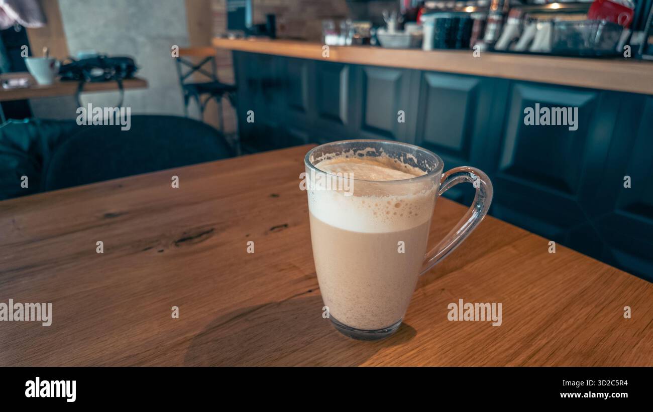 Glass cup of coffee on wooden table near window inside urban cafe, morning light creating warm atmosphere perfect for lifestyle or business concepts Stock Photo