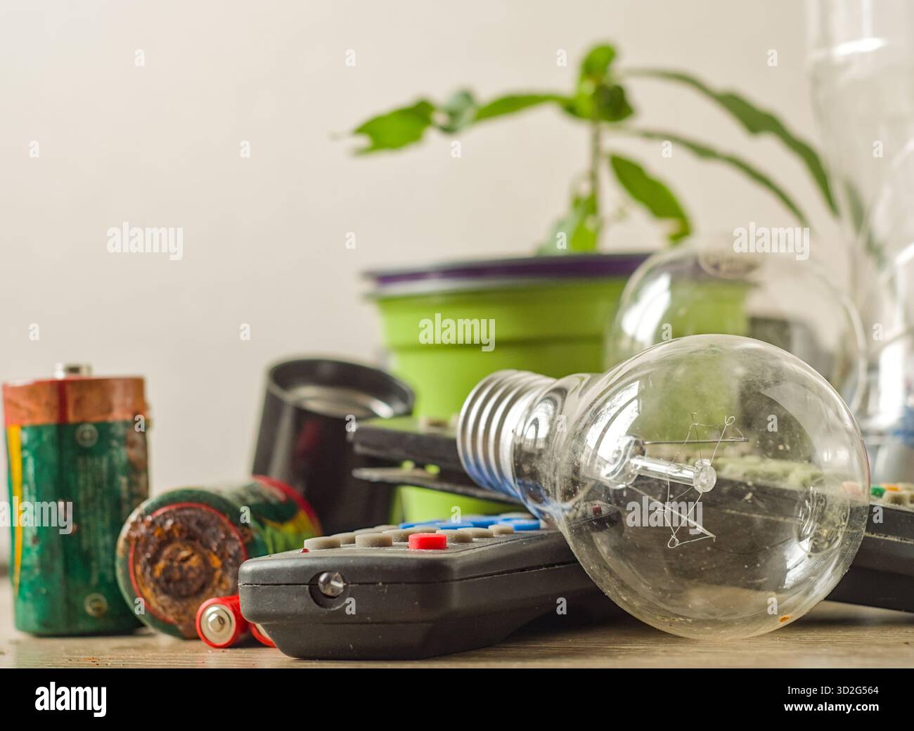 Old incandescent light bulb, used remote controls and old batteries, grouped around a small houseplant to symbolize the challenge of e-waste recycling Stock Photo
