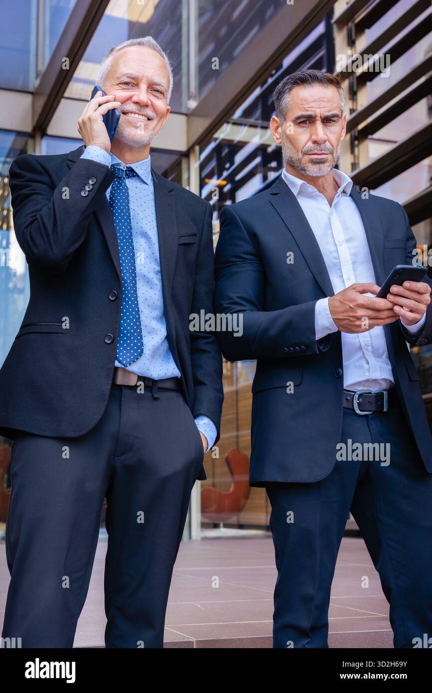 Two businessmen in suits using smartphones, talking outside modern ...