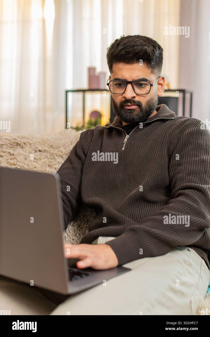 Indian man at home types on laptop with focused expression, preparing for upcoming online exam with full attention. Arabian guy on sofa concentrates on studying and memorizing materials for success Stock Photo