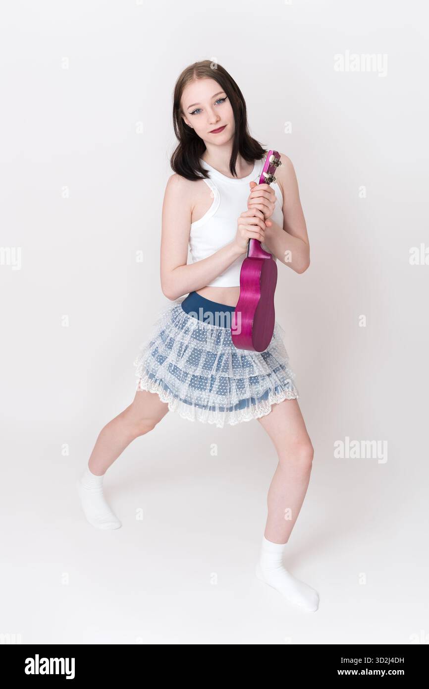 Skinny woman dancer and musician is standing and holding small guitar called ukulele. Eighteen year old woman is wearing white singlet, short skirt an Stock Photo