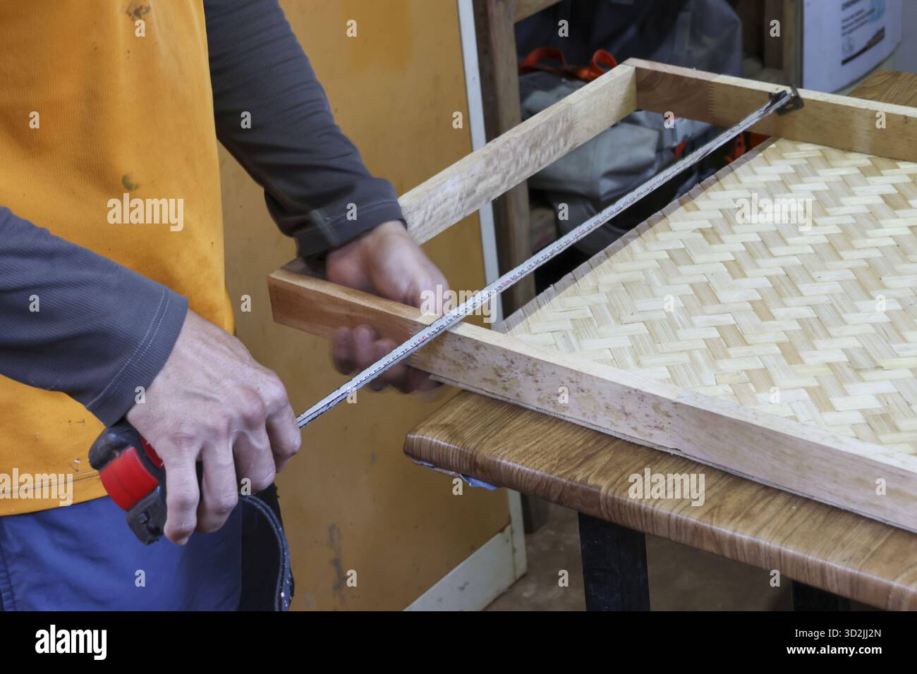 Focused craftsman measuring handmade wooden frame with woven bamboo bottom in workshop. This carpenter uses tape measure for precise DIY furniture pro Stock Photo