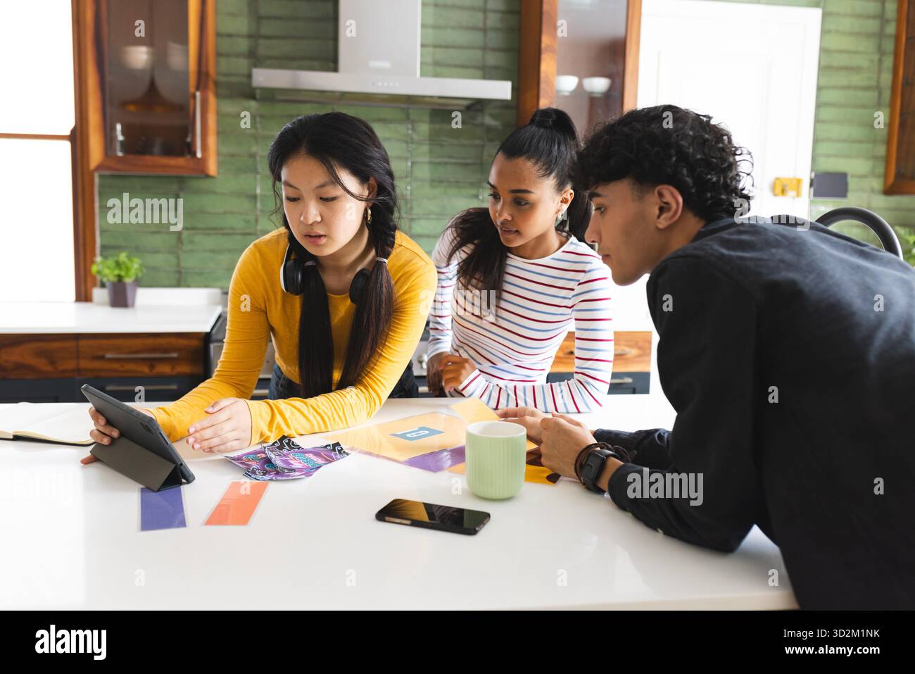 Teenagers collaborating on project using tablet in modern kitchen setting. Collaboration, teamwork, technology, brainstorming, creativity, discovery || Model released Stock Photo