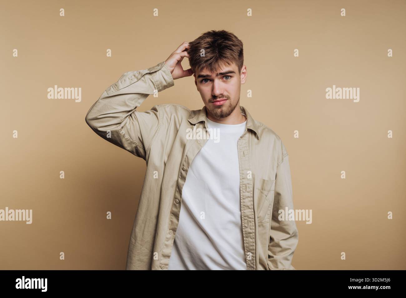 Young man in casual clothing scratching his head, beige background. Stock Photo
