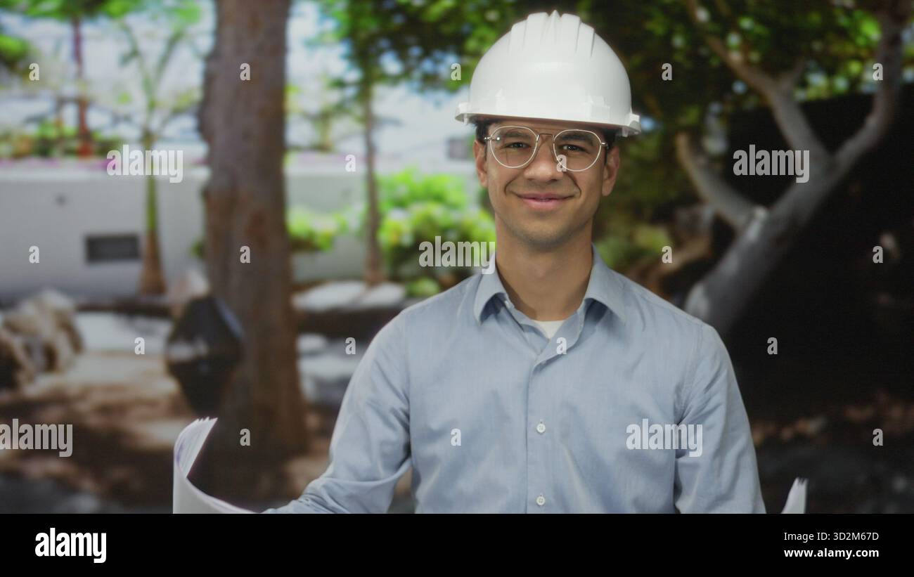 Young hispanic man wearing hardhat reviews blueprints while smiling near building; planning confidence. Stock Photo