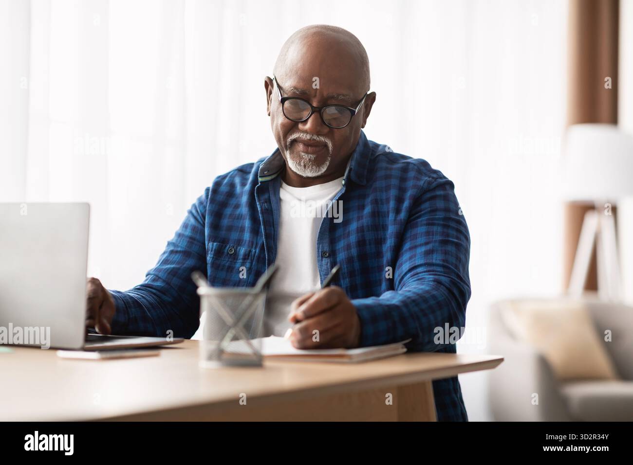 Mature African American Man Taking Notes While Using Laptop in Modern Office Environment Stock Photo