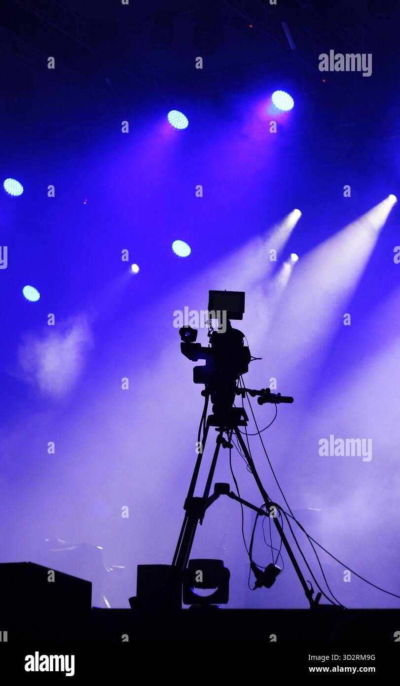 Silhouette of a professional video camera on a tripod next to a stage with dramatic blue and purple lighting Stock Photo