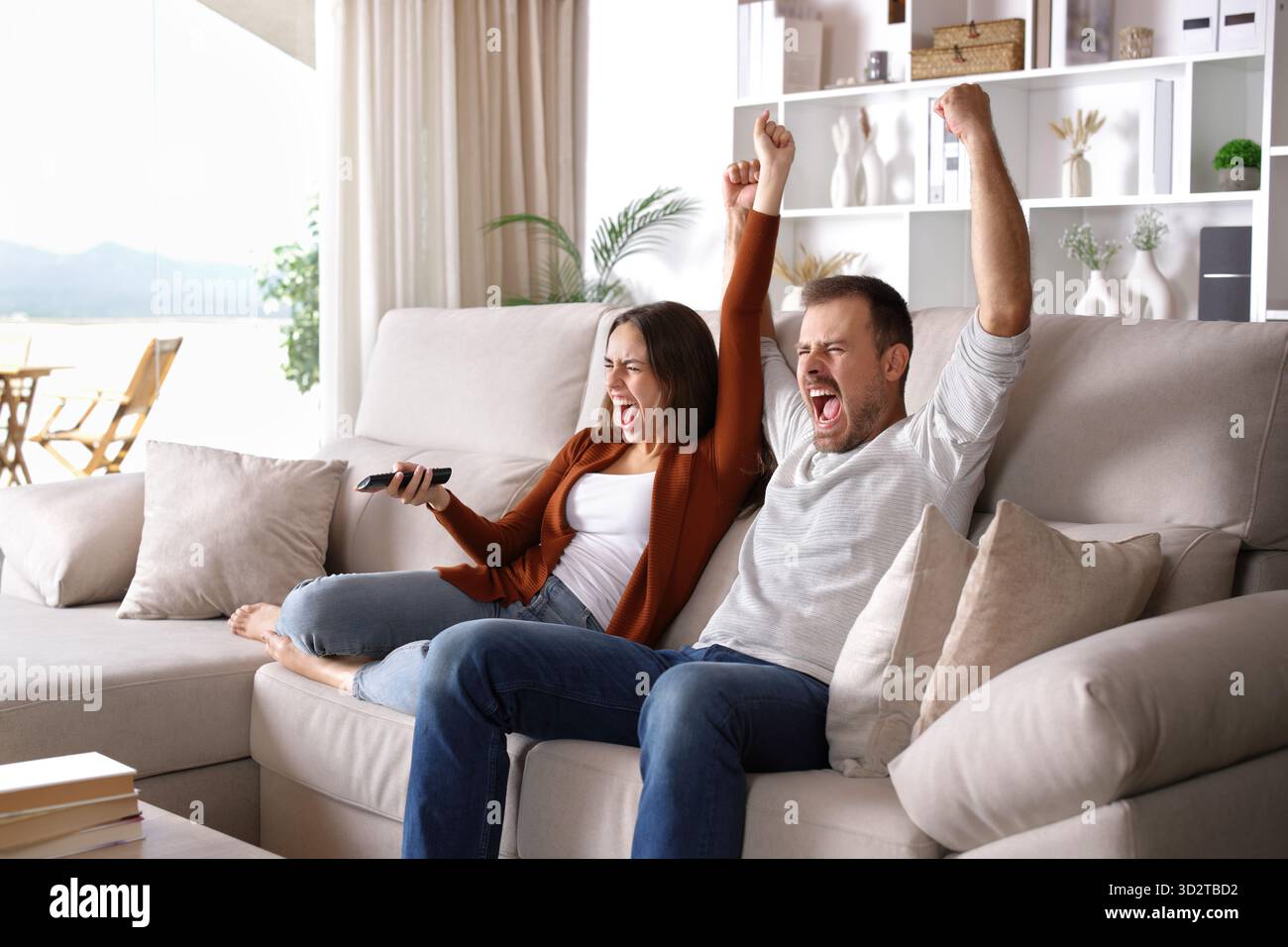 Excited couple watching tv celebrating sitting on a couch at home Stock Photo