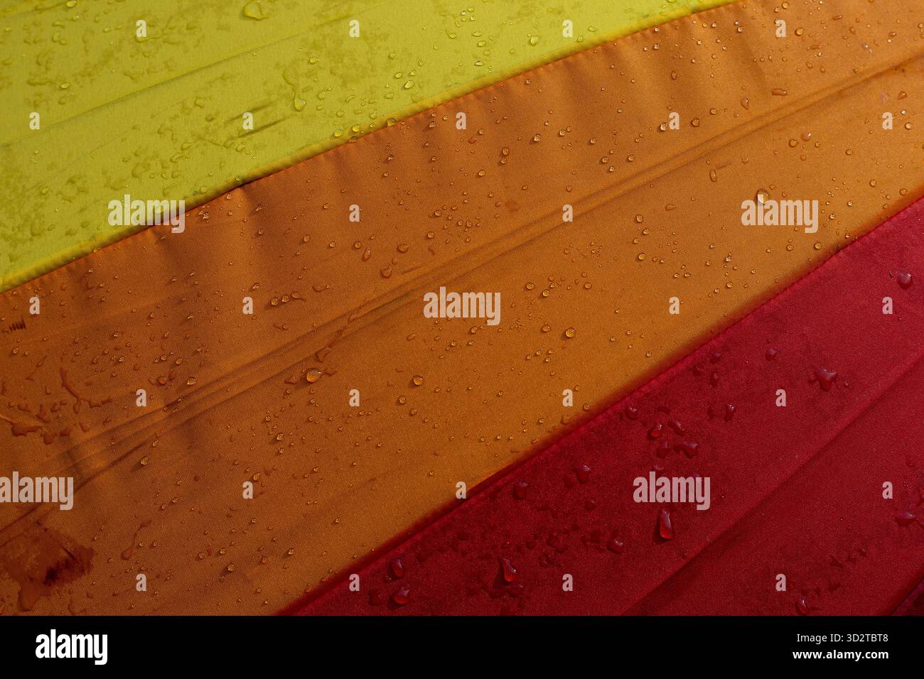Colorful umbrella fabric with vibrant yellow, orange, and red stripes appears wet with numerous small water droplets. abstract close up creates refres Stock Photo