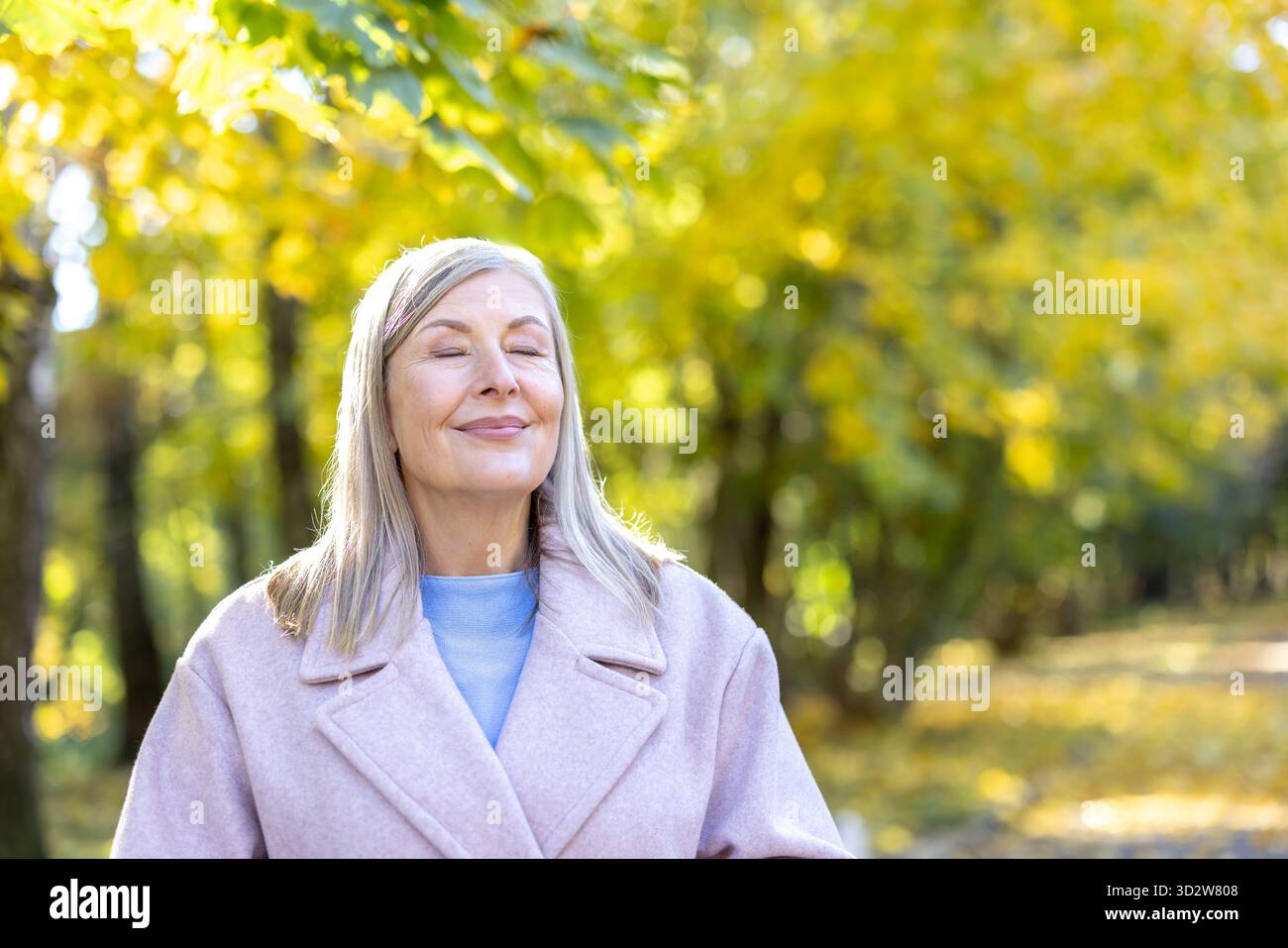 Senior woman relaxing with eyes closed and a gentle smile, feeling the sun on her face in an autumn park with blurred yellow foliage in the background Stock Photo