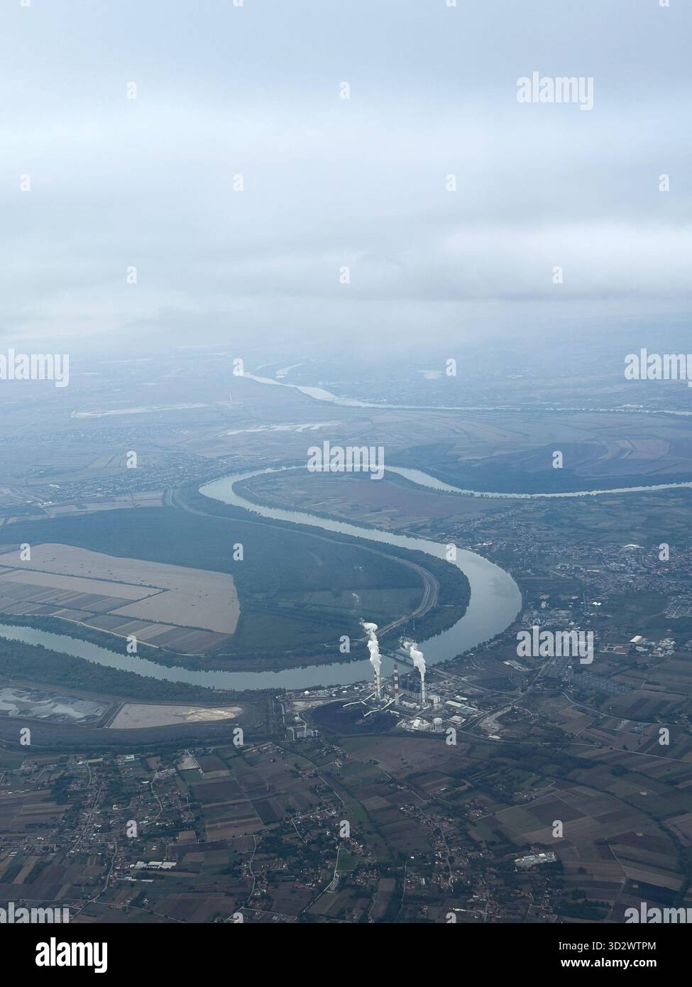 Scenic aerial view of a winding river flowing through flat farmland with an industrial plant emitting steam under a cloudy sky. - Smartphone Captured Stock Image