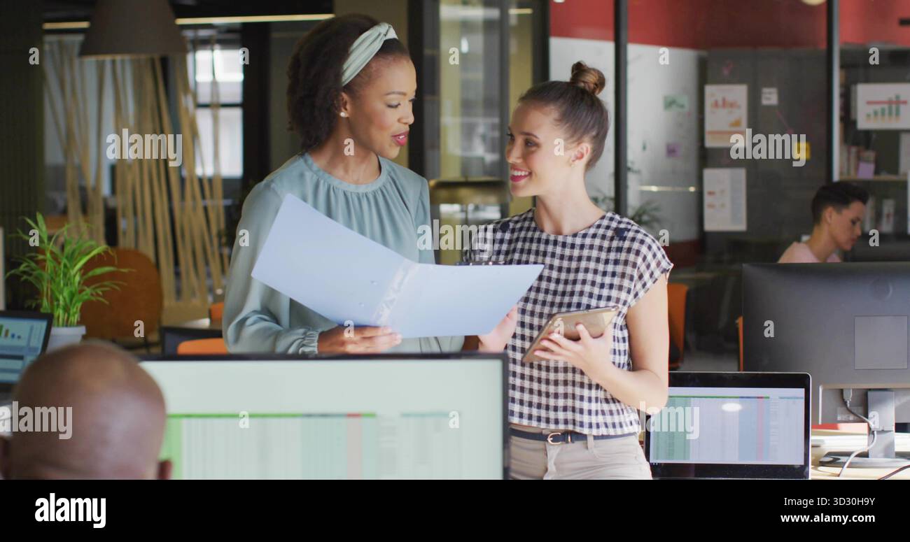 Reviewing project documents two women collaborating in open-plan office, with folder and tablet Stock Photo