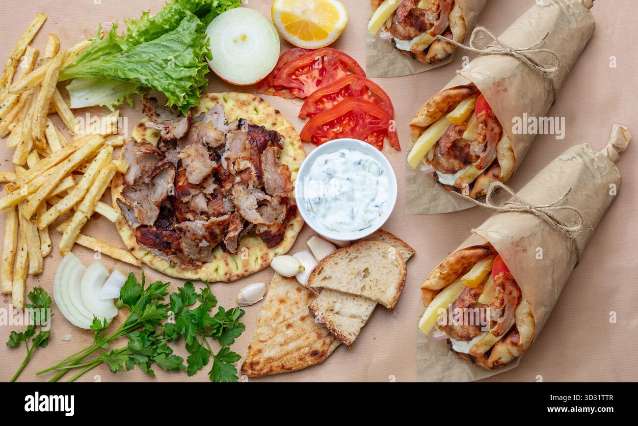 Greek gyros wraps and fresh ingredients arranged on a rustic table under natural daylight, showing pita bread, grilled meat, fries, vegetables, and tz Stock Photo
