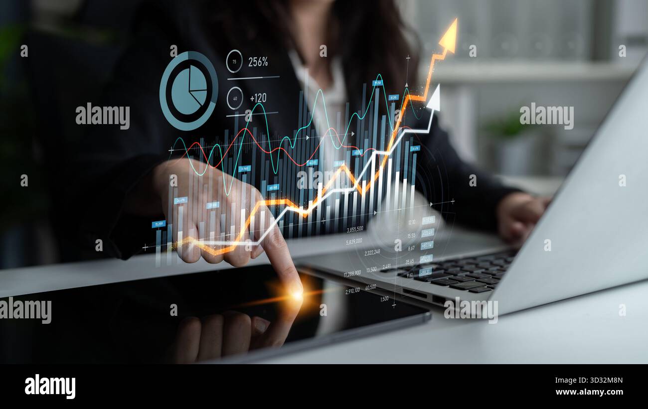 A businesswoman is analyzing financial data with digital graphs on her laptop and interacting with a tablet in a modern office, showcasing strategic i Stock Photo