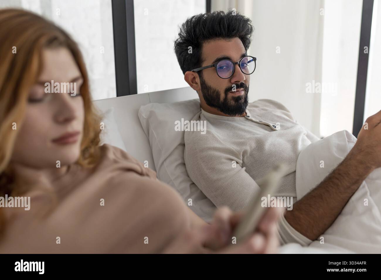 Young Mixed-race couple with serious expression using smartphone lying on bed Stock Photo