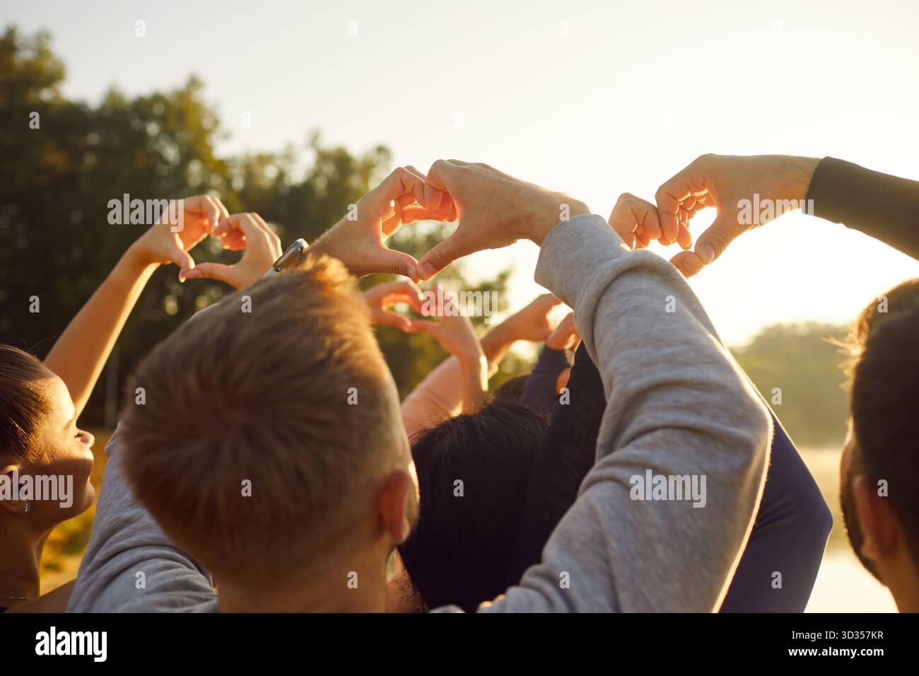 People making heart shape gesture with fingers in sunshine, back view of happy young friends crowd Stock Photo