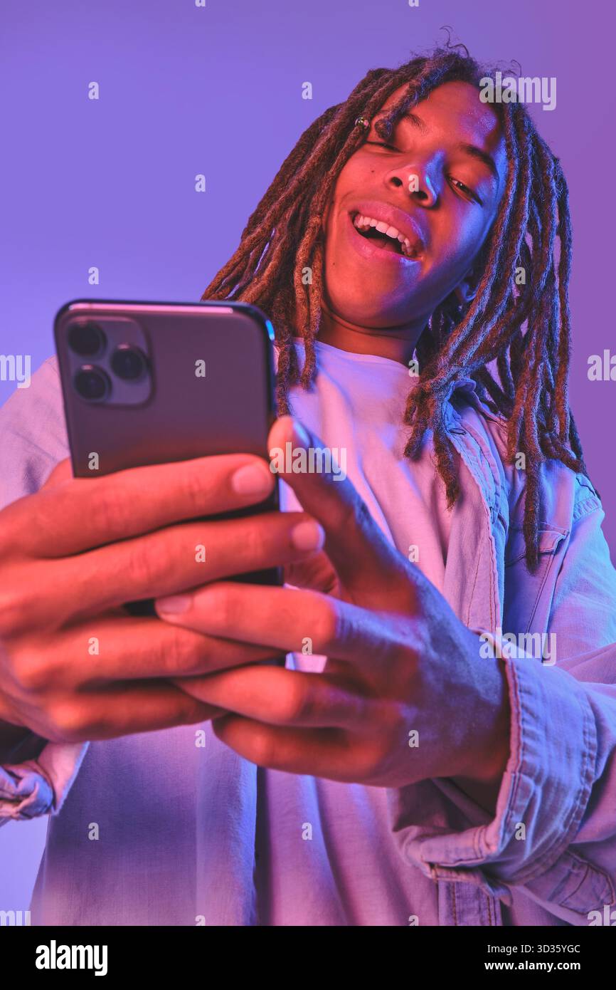 Young man with dreadlocks smiling while holding and using a smartphone Stock Photo