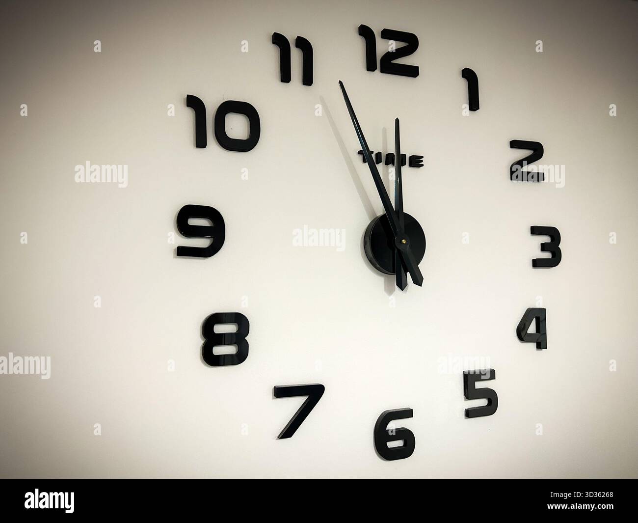 Minimalist wall clock with black numbers and hands showing 11:55 on a white background, symbolizing urgency, deadlines, and time running out. - Smartphone Captured Stock Image