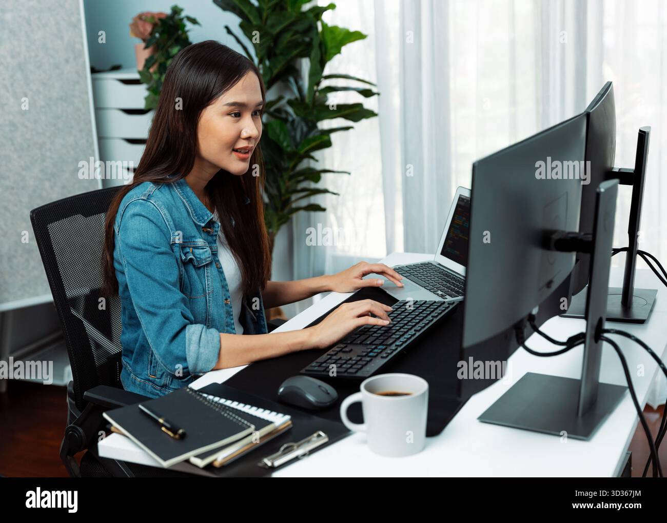 Smiling young Asian IT developer working software development information on pc screens and laptop, creating program coding for latest version Stock Photo