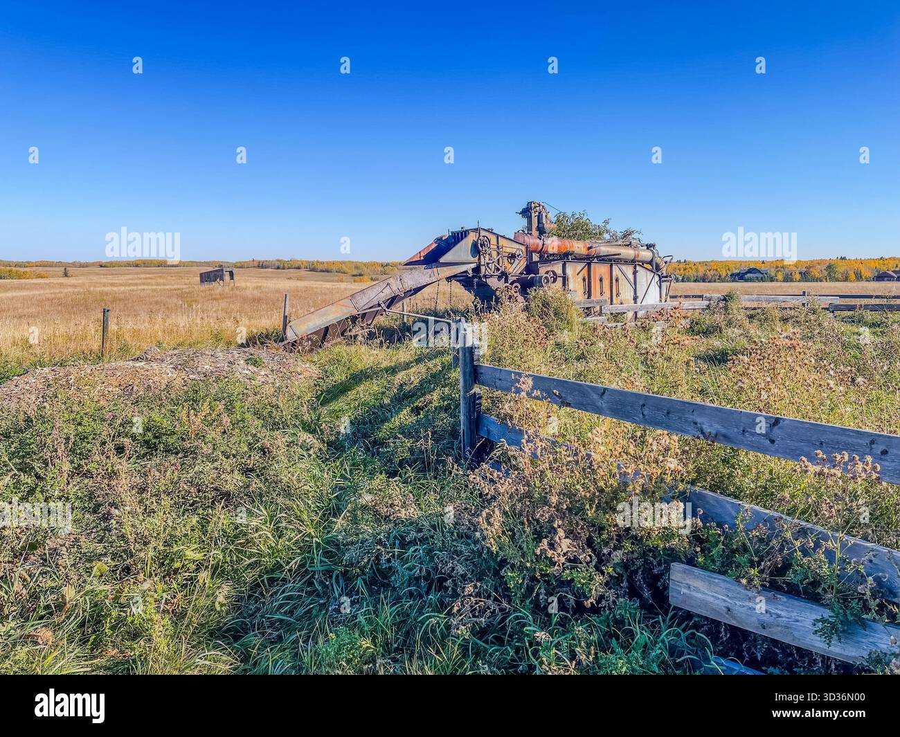 Rusty, abandoned machinery sits in a wide rural field beside a weathered wooden fence. The scene features a calm sky, open landscape, and overgrown ve - Smartphone Captured Stock Image