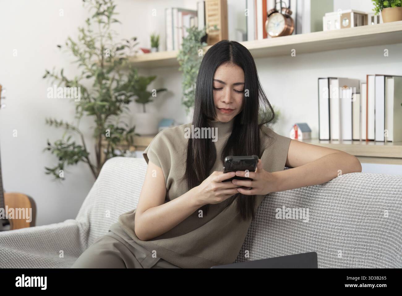 A focused young woman engages with her smartphone while sitting comfortably on a couch in a cozy home environment Stock Photo