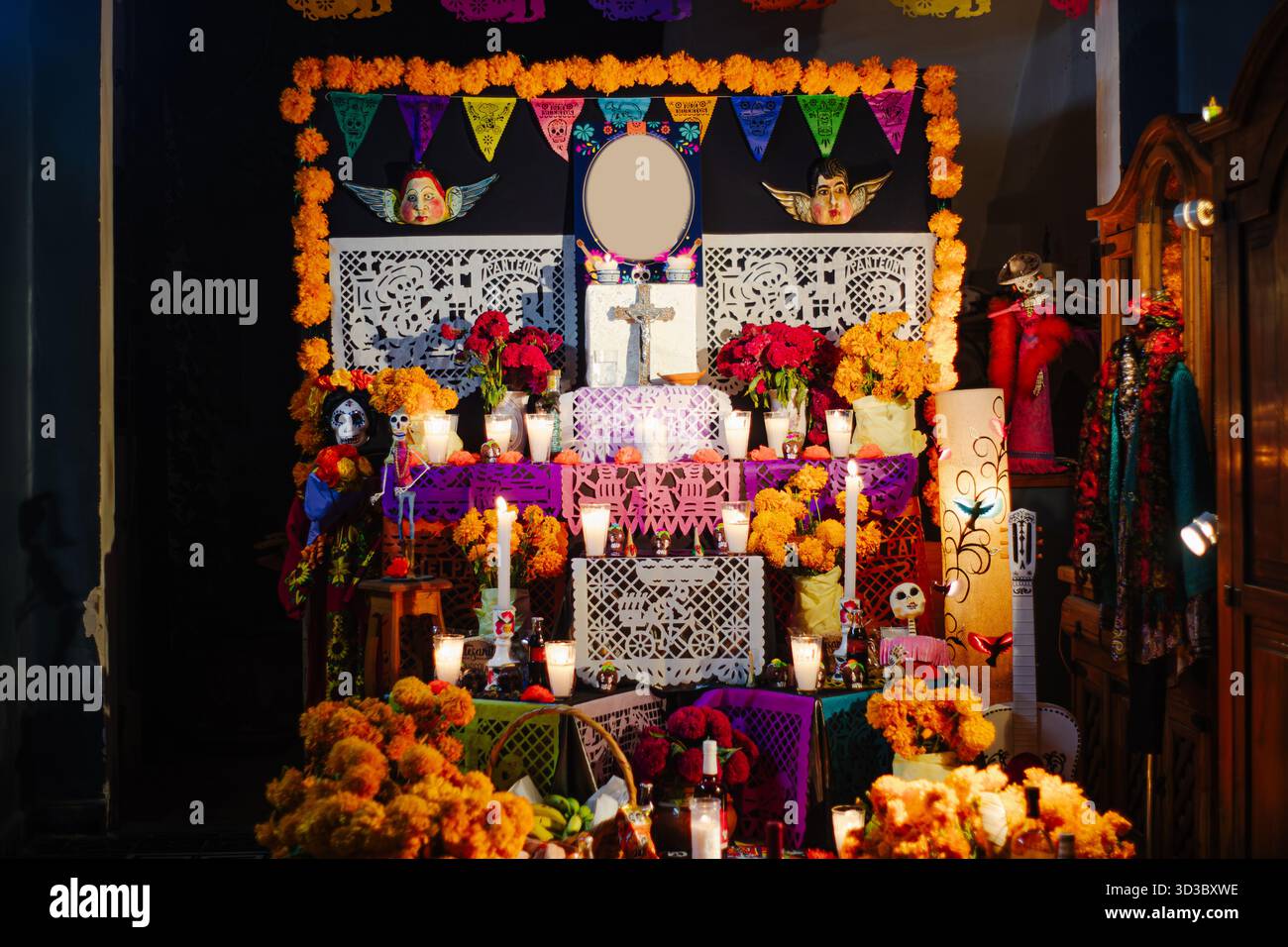 Front view of a richly decorated Day of the Dead altar with roses, marigold, white cutout papel picado, and lit candles against a dark background. Stock Photo