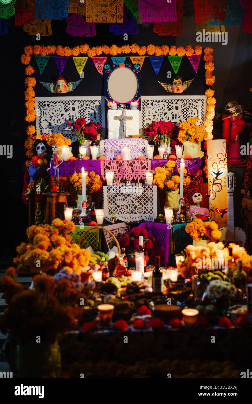 Front view of a richly decorated Day of the Dead altar with roses, marigold, white cutout papel picado, and lit candles against a dark background. Stock Photo