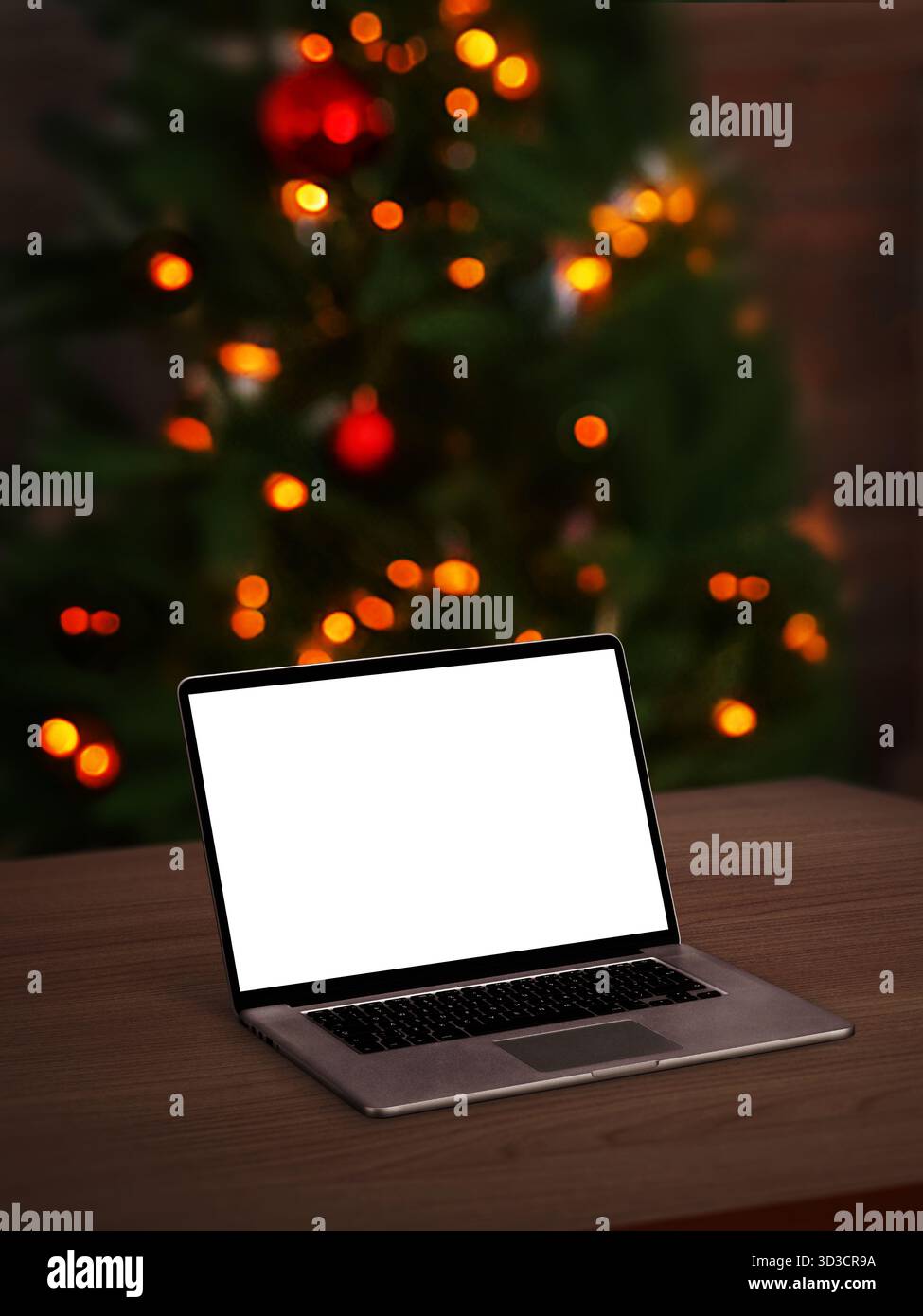 Open laptop with blank screen on wooden table, Christmas tree lights glowing in soft focus background, cozy festive atmosphere Stock Photo