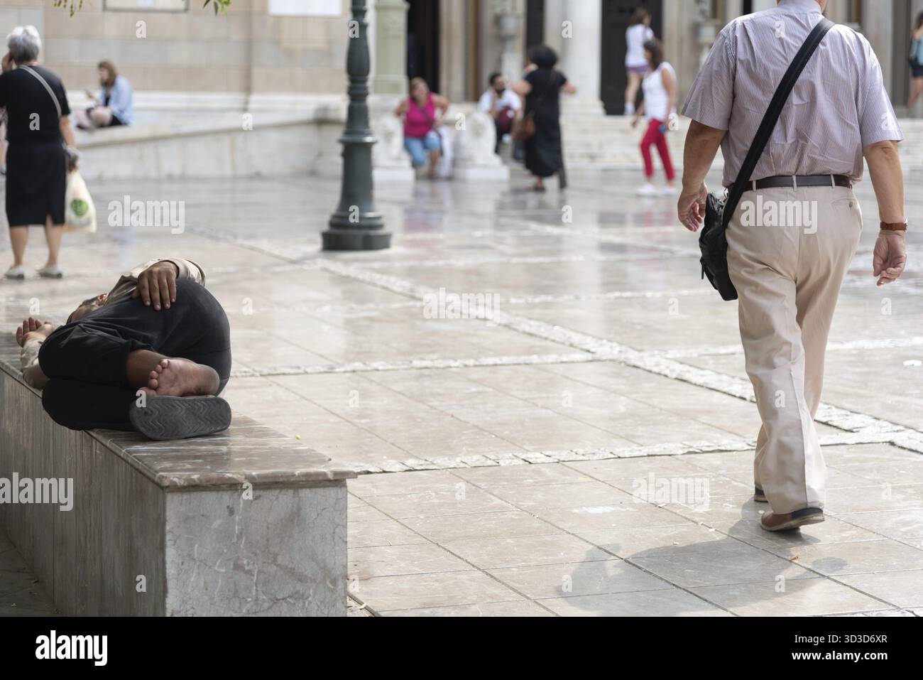 Homeless man sleeping on a bench in a city square while a tourist walks ...