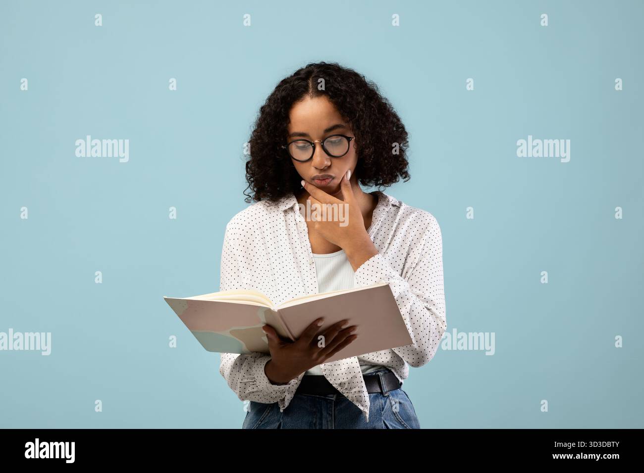 Pensive Black Female Student Reading Textbook While Studying for Exam Against Blue Background Stock Photo