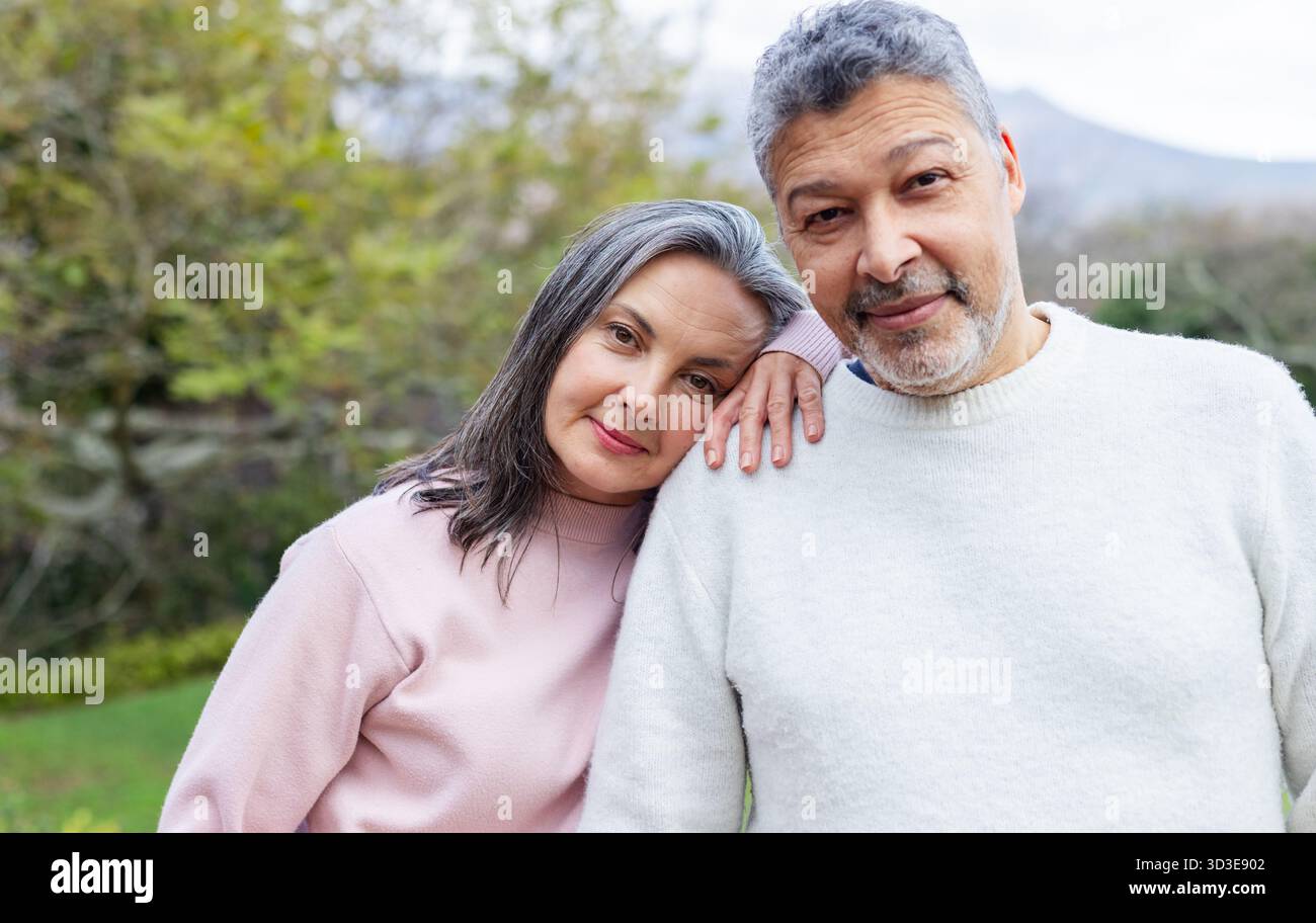 Senior couple in sweaters standing close together looking at camera in grassy yard under cloudy sky Stock Photo