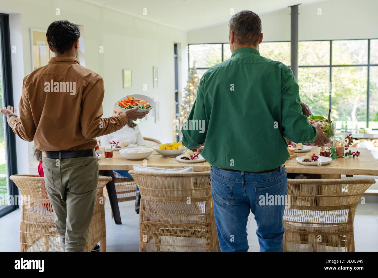 Diverse family carrying large salad bowls toward seated members at wooden dining table by windows Stock Photo