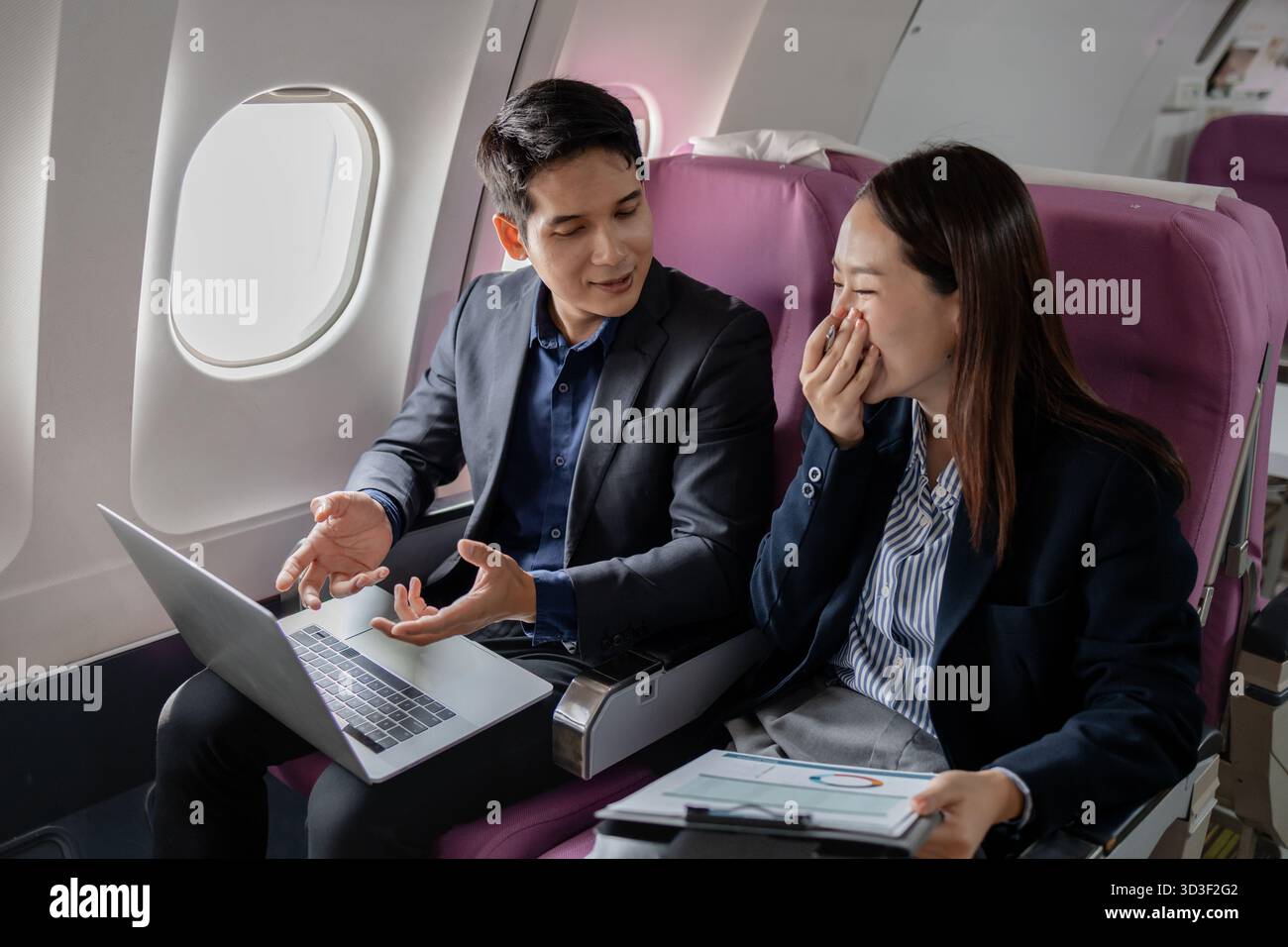 Two Asian business professionals in formal suits discussing work mid-flight. Seated on an airplane, they are exchanging ideas with expressive hand ges Stock Photo