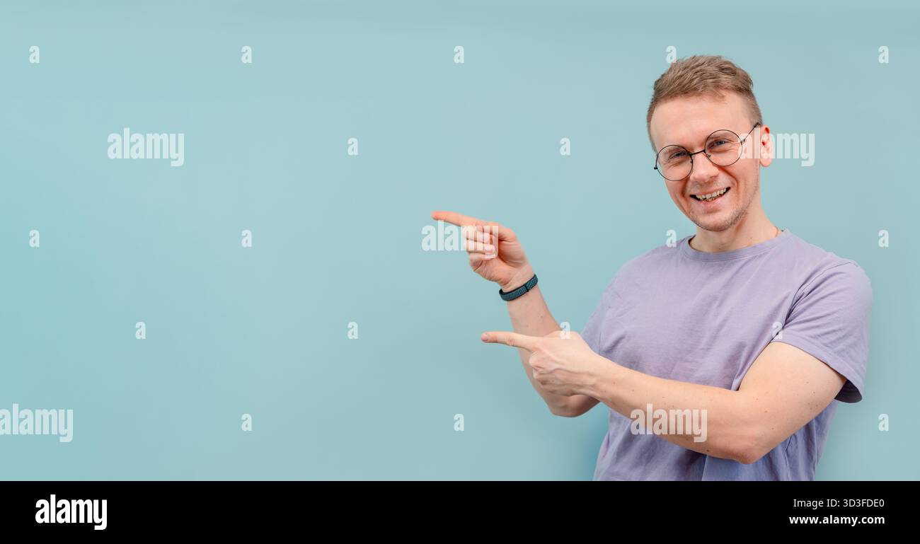 A cute funny young man in nerdy glasses points with his fingers at the blue background for the banner and text Stock Photo
