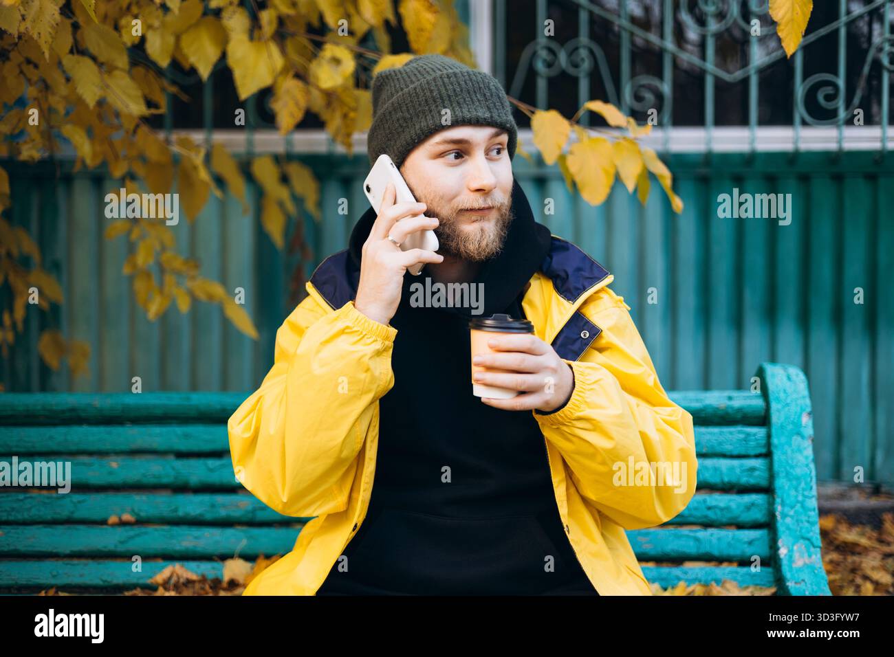 Man wearing a bright yellow raincoat sits on a bench surrounded by autumn leaves, holding a cup of coffee and talking on the phone, embracing the fall Stock Photo