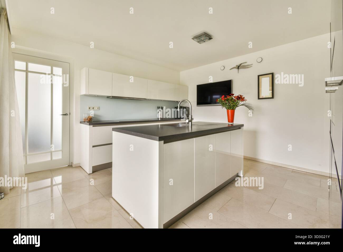 A stylish modern kitchen featuring sleek white cabinetry, elegant countertops, and a vibrant floral centerpiece, creating a bright and inviting atmosp Stock Photo