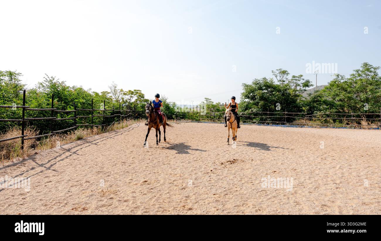 Two girls ride horses in an outdoor training arena while both are using ...