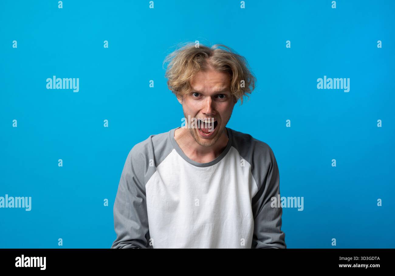 Angry man in gray and white shirt shouting with open mouth on blue background Stock Photo