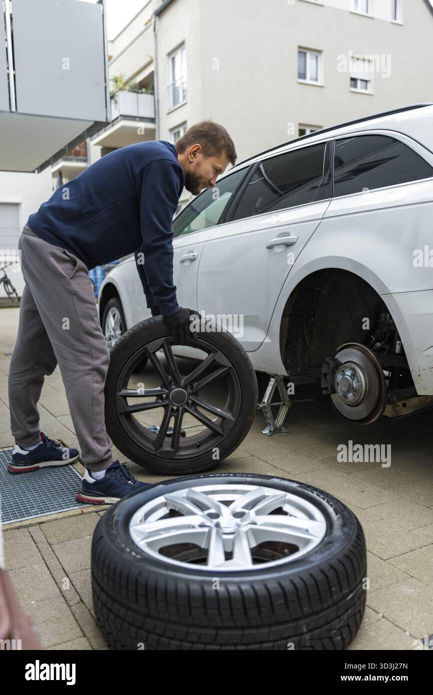 Man changing car tire using electric drill and jack on city sidewalk. DIY auto repair, maintenance, seasonal tire change, mechanical work, vehicle Stock Photo