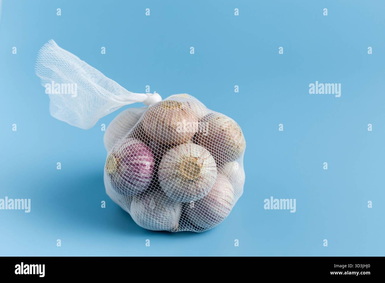 Small garlic bulbs in a mesh bag on a solid blue background. Stock Photo