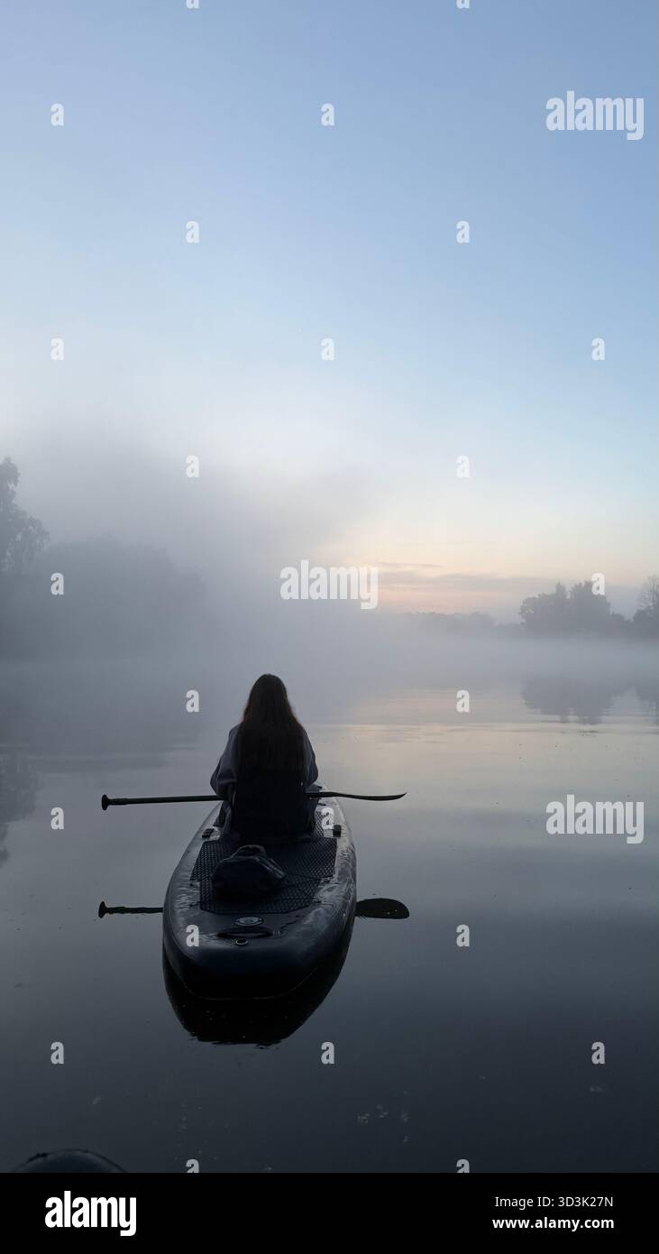 A woman sitting on a paddleboard faces the misty river ahead, surrounded by dense fog and a calm, tranquil atmosphere - Smartphone Captured Stock Image