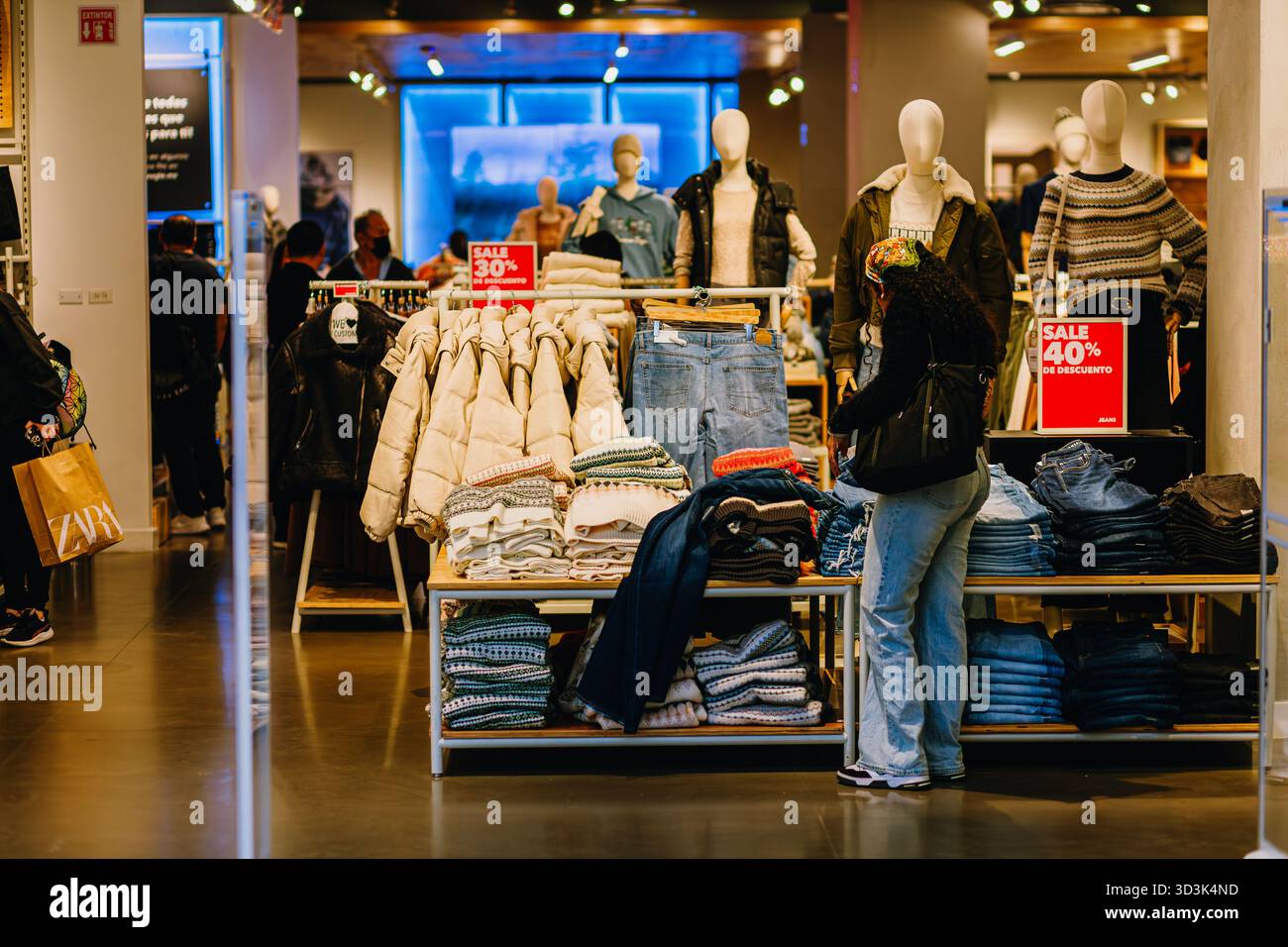 Female customer in a casual clothing store examining jeans and coats with 30% and 40% sale promotion. Mannequins in the background. Stock Photo