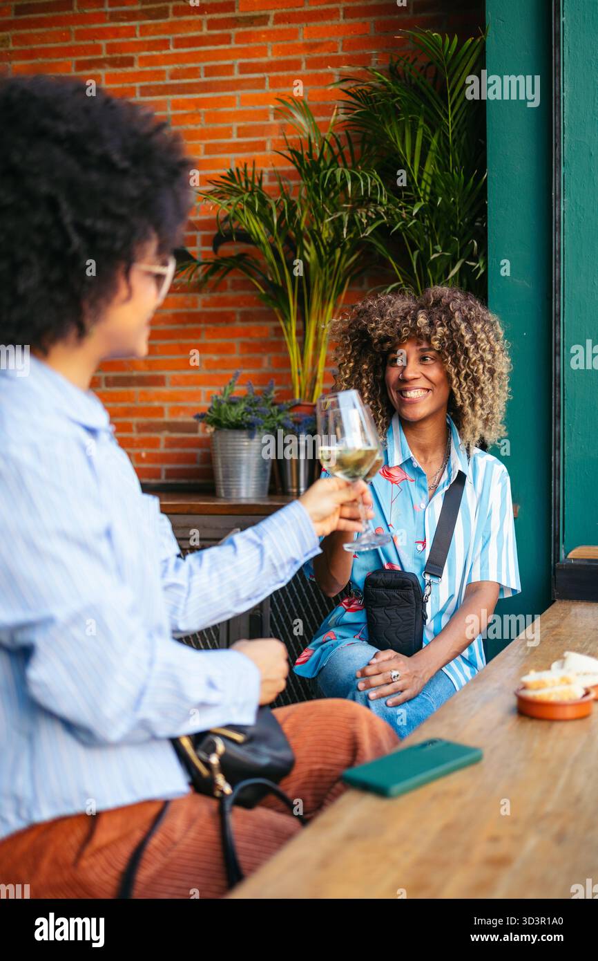 Two friends share a moment over drinks in a lively downtown setting. Surrounded by vibrant plants and warm brick walls, they enjoy a sunny day filled Stock Photo