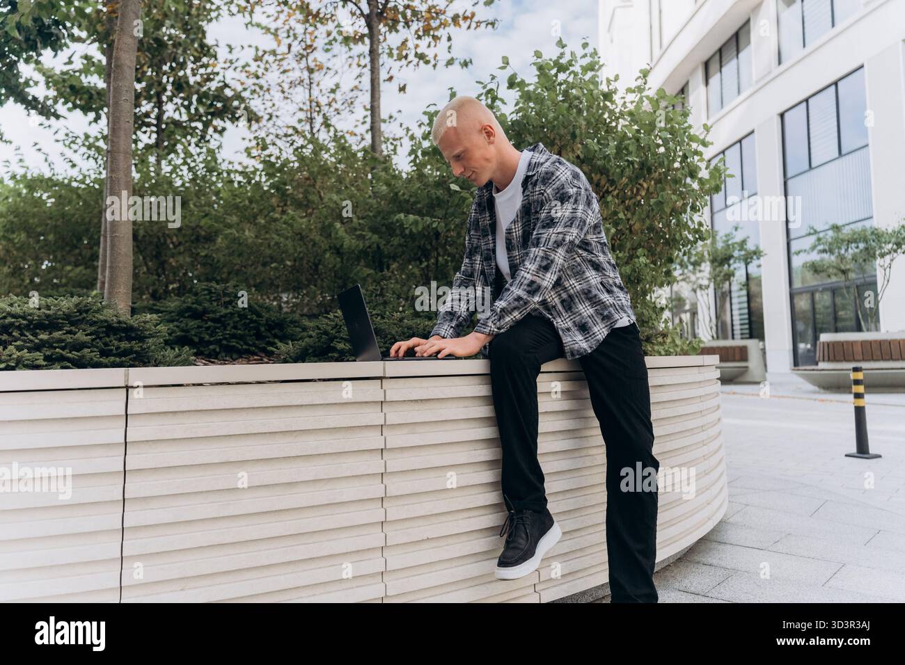 A young man with an albino appearance is focused on his laptop while leaning on a modern, urban planter in a city environment, exuding concentration a Stock Photo