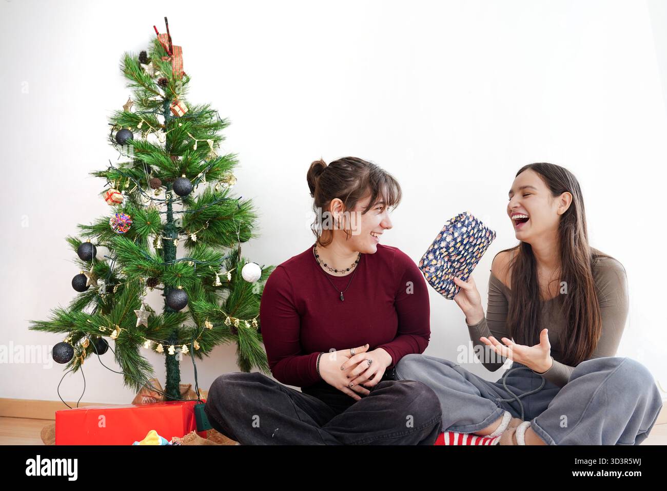 Two girls share laughter and gifts by a beautifully decorated Christmas tree in a cozy home setting. Festive winter spirit is in the air as they enjoy Stock Photo
