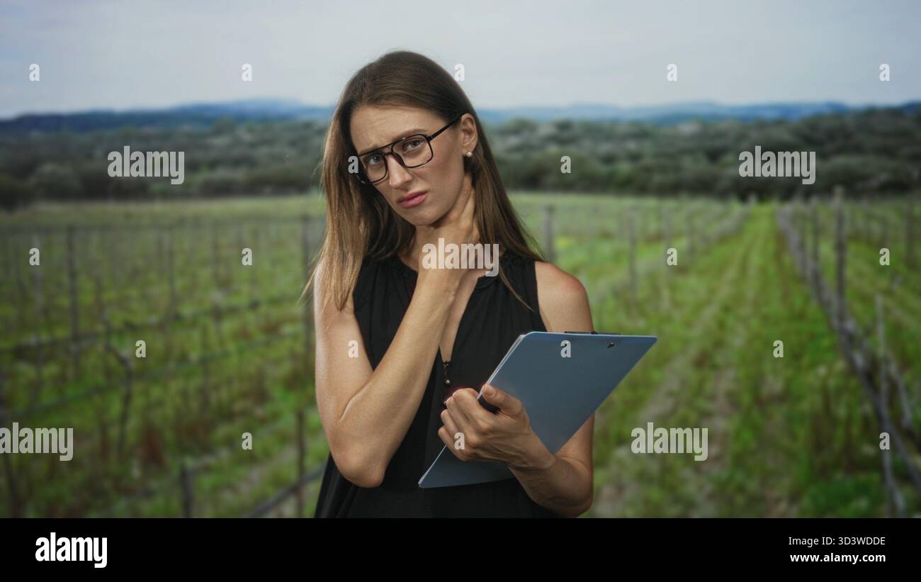 Woman holding clipboard and touching her neck while frowning in a forest vineyard field inspection; rural inspection concern. Stock Photo