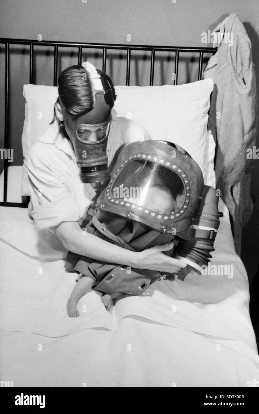 A mother and baby both in gas-masks during 1941. A mother cradles her newborn baby in bed, shortly after giving birth. The mother is wearing her civilian respirator, whilst the baby is encased in a baby gas helmet, which buckles up around the baby's bottom. The mother is demonstrating how the bellows on her baby's gas mask are pumped to supply the baby with air. Stock Photo